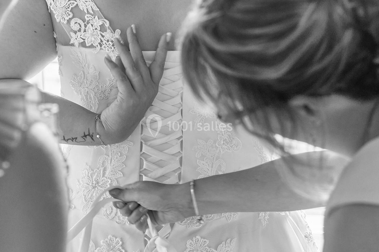 Une mariée en robe blanche se fait ajuster le laçage du dos par deux personnes.