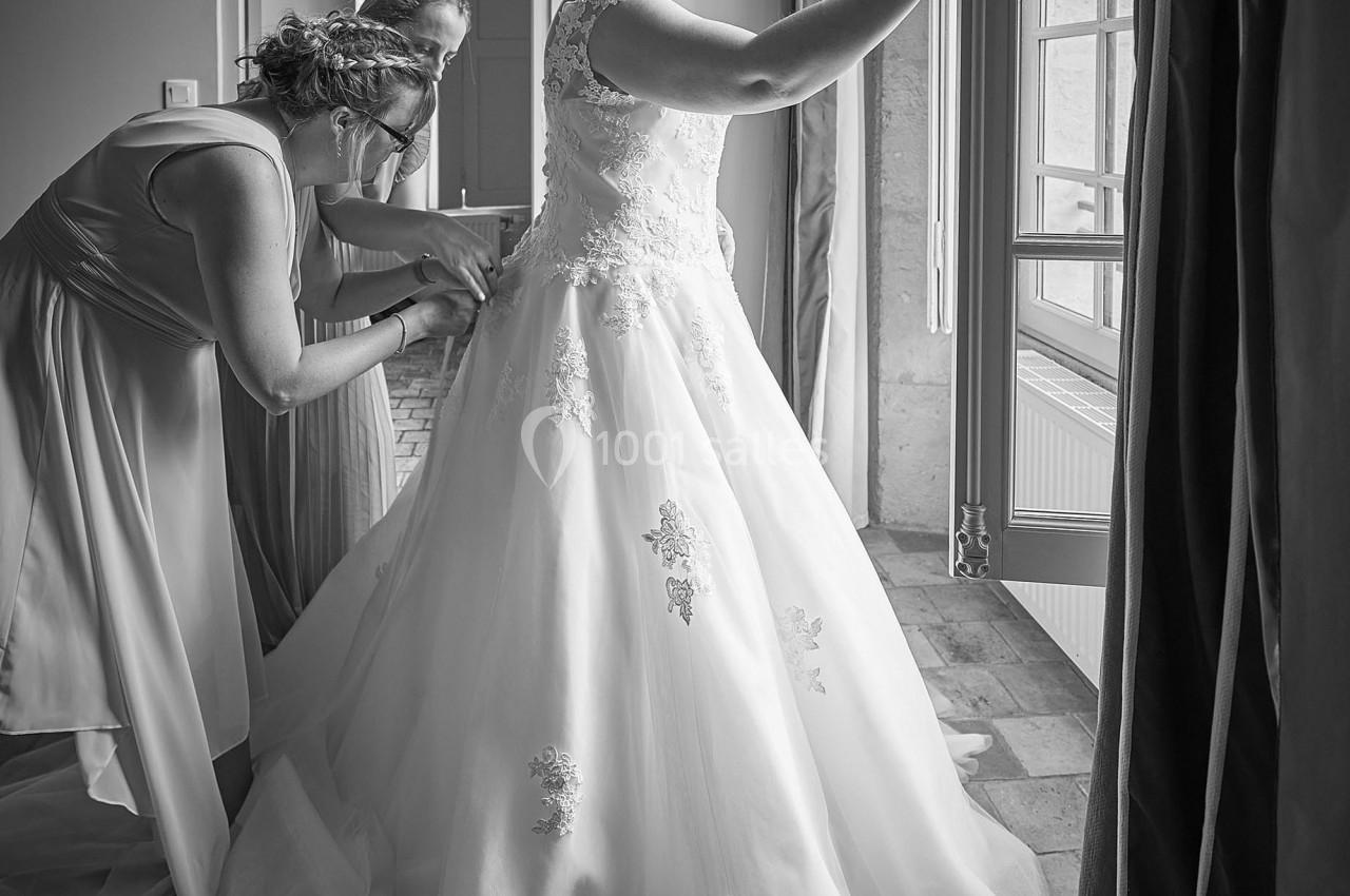 Une mariée en robe blanche regarde par la fenêtre tandis que deux personnes ajustent sa tenue.