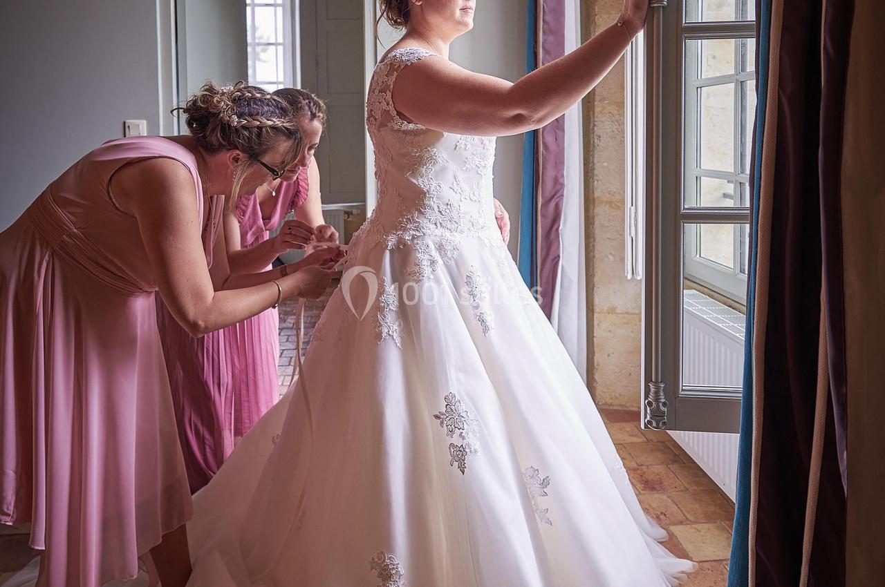 Une mariée en robe blanche regarde par une fenêtre tandis qu'une femme ajuste sa tenue dans une pièce lumineuse.