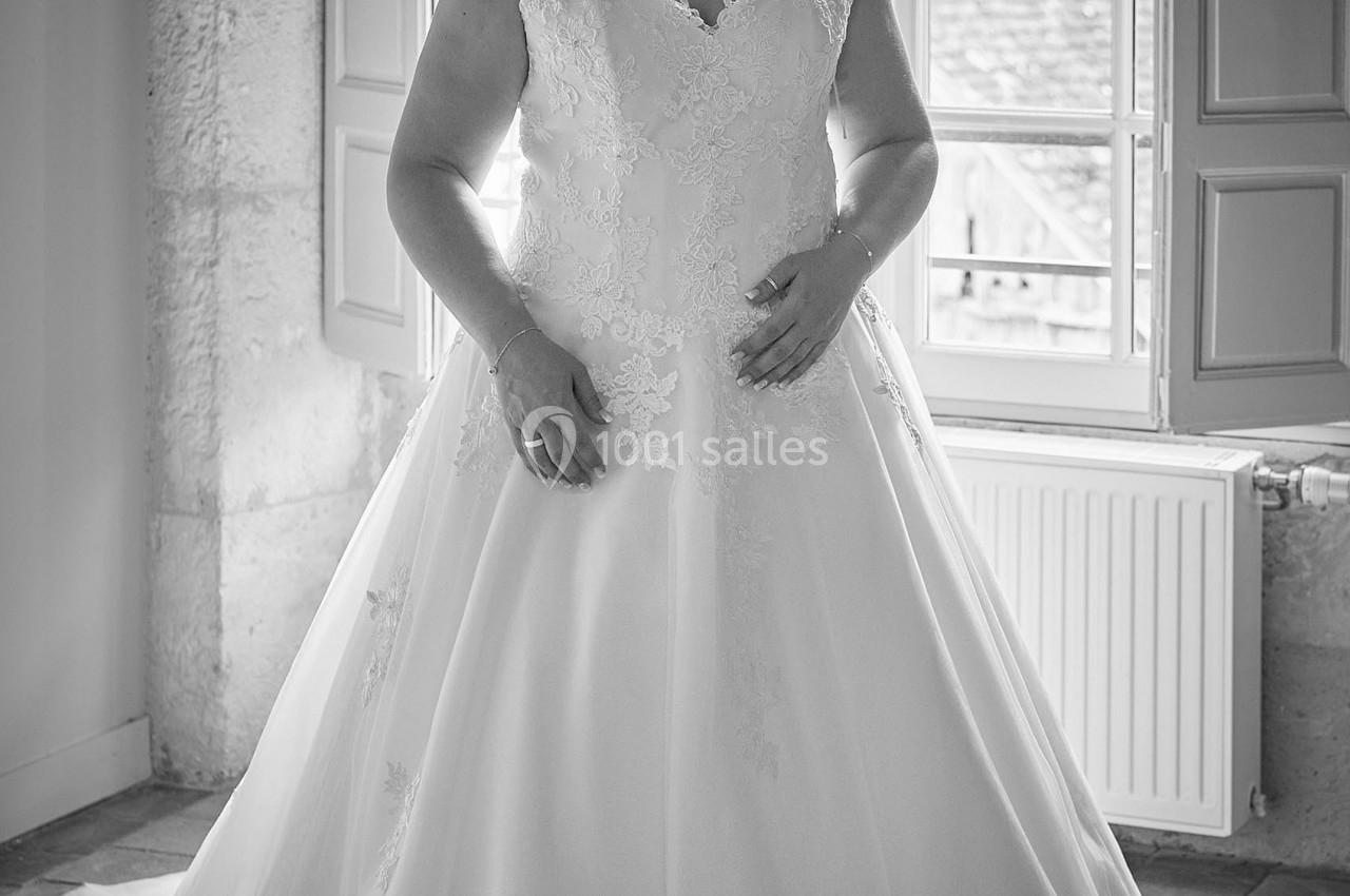 Femme en robe de mariée élégante, debout près d'une fenêtre ouverte laissant entrer la lumière naturelle.