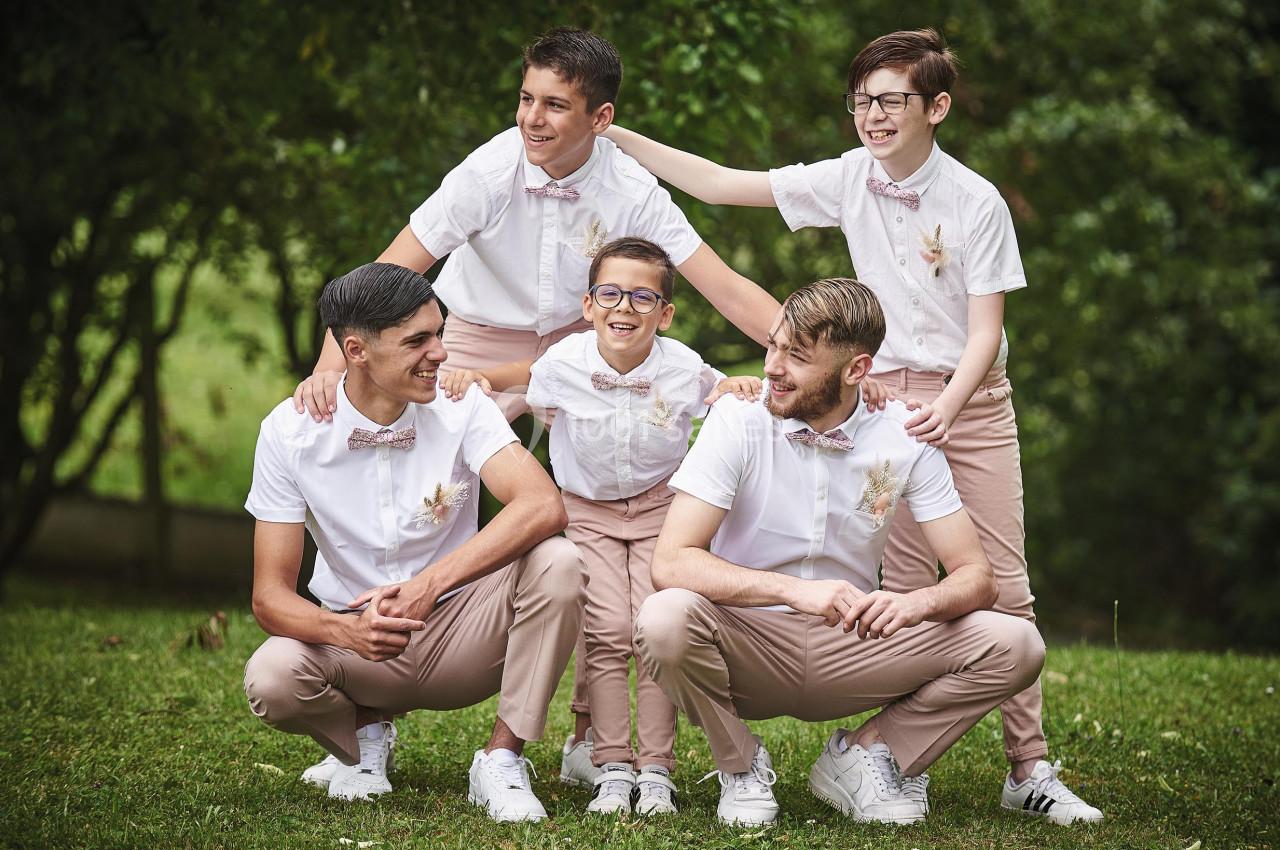 Cinq garçons en tenue assortie, chemises blanches et pantalons beiges, posent souriants dans un parc verdoyant.