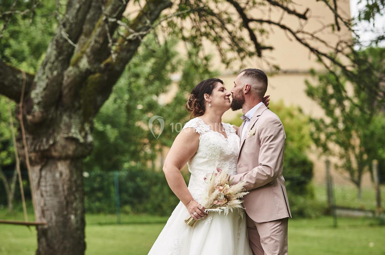 Un couple en tenue de mariage pose sous un arbre dans un jardin verdoyant, l'homme embrassant la femme sur le front.