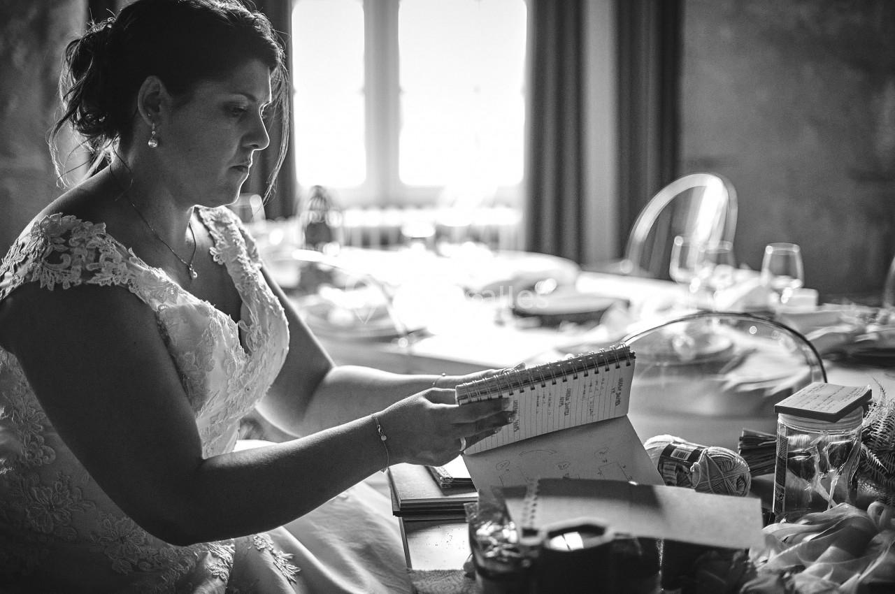 Une femme en robe de mariée lit un carnet dans une pièce éclairée par une fenêtre en arrière-plan.
