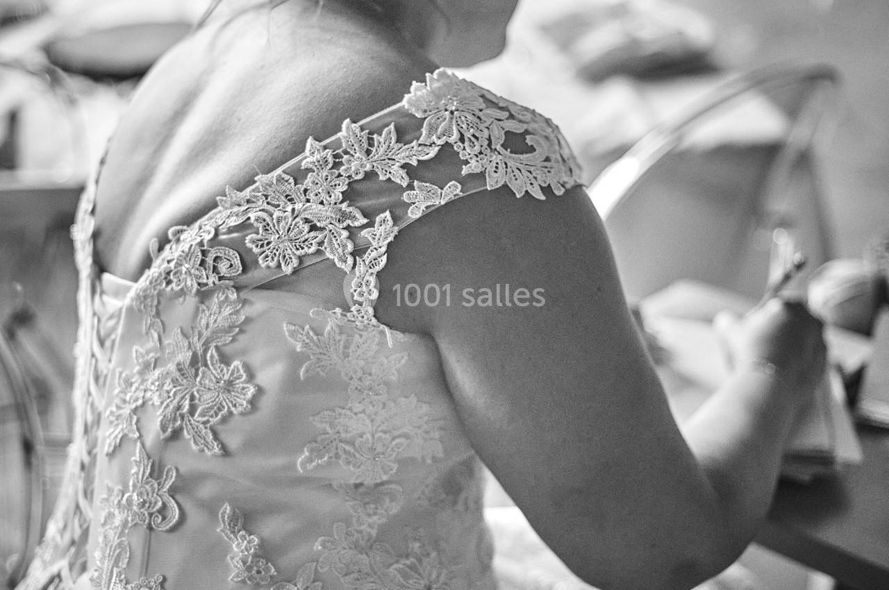 Femme en robe de mariée brodée, assise de dos, écrivant sur une table dans une pièce éclairée naturellement.