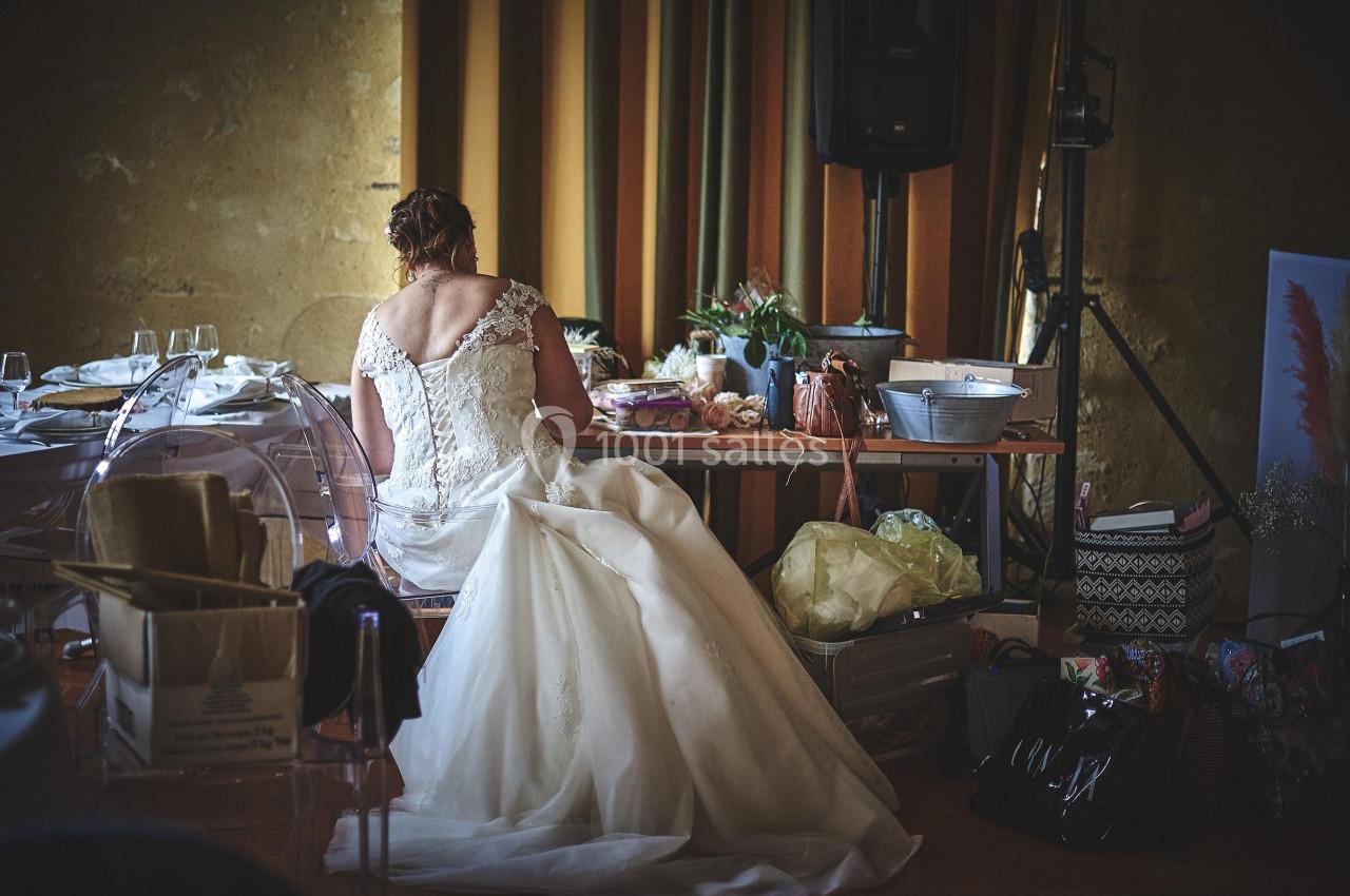 Une femme en robe de mariée est assise de dos devant une table encombrée dans une salle décorée.