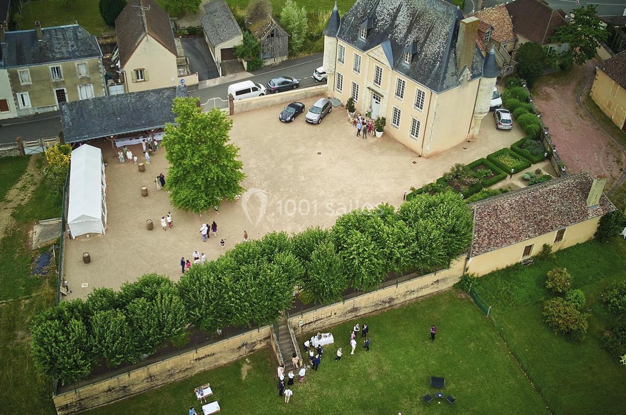 Vue aérienne d'un manoir entouré de jardins, avec des invités rassemblés dans la cour et sur la pelouse.