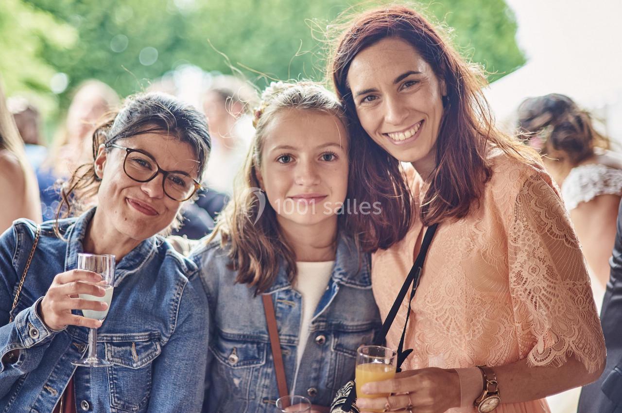 Trois femmes souriantes, dont une adolescente, posent ensemble en extérieur, tenant des verres à la main.