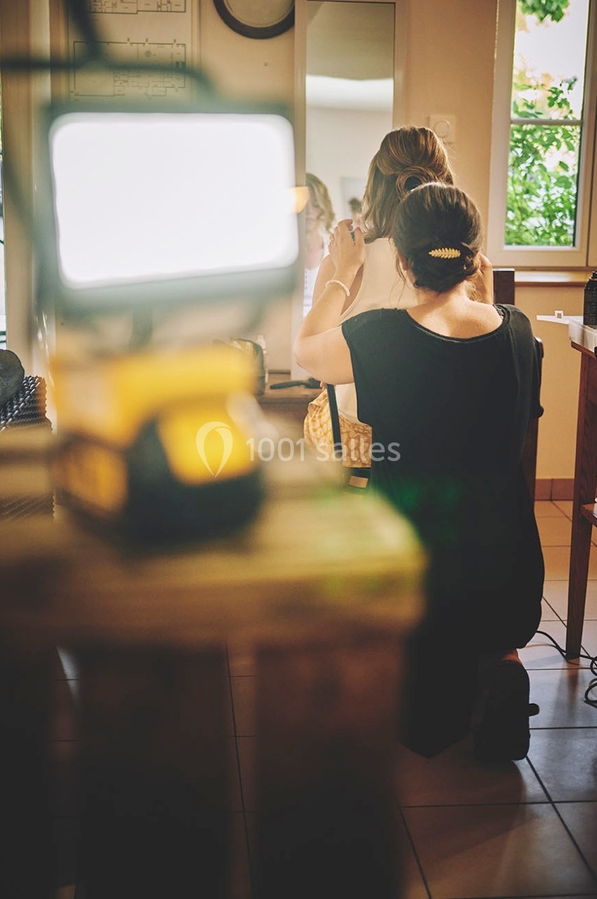 Une coiffeuse ajuste la coiffure d'une femme assise devant un miroir dans une pièce lumineuse.