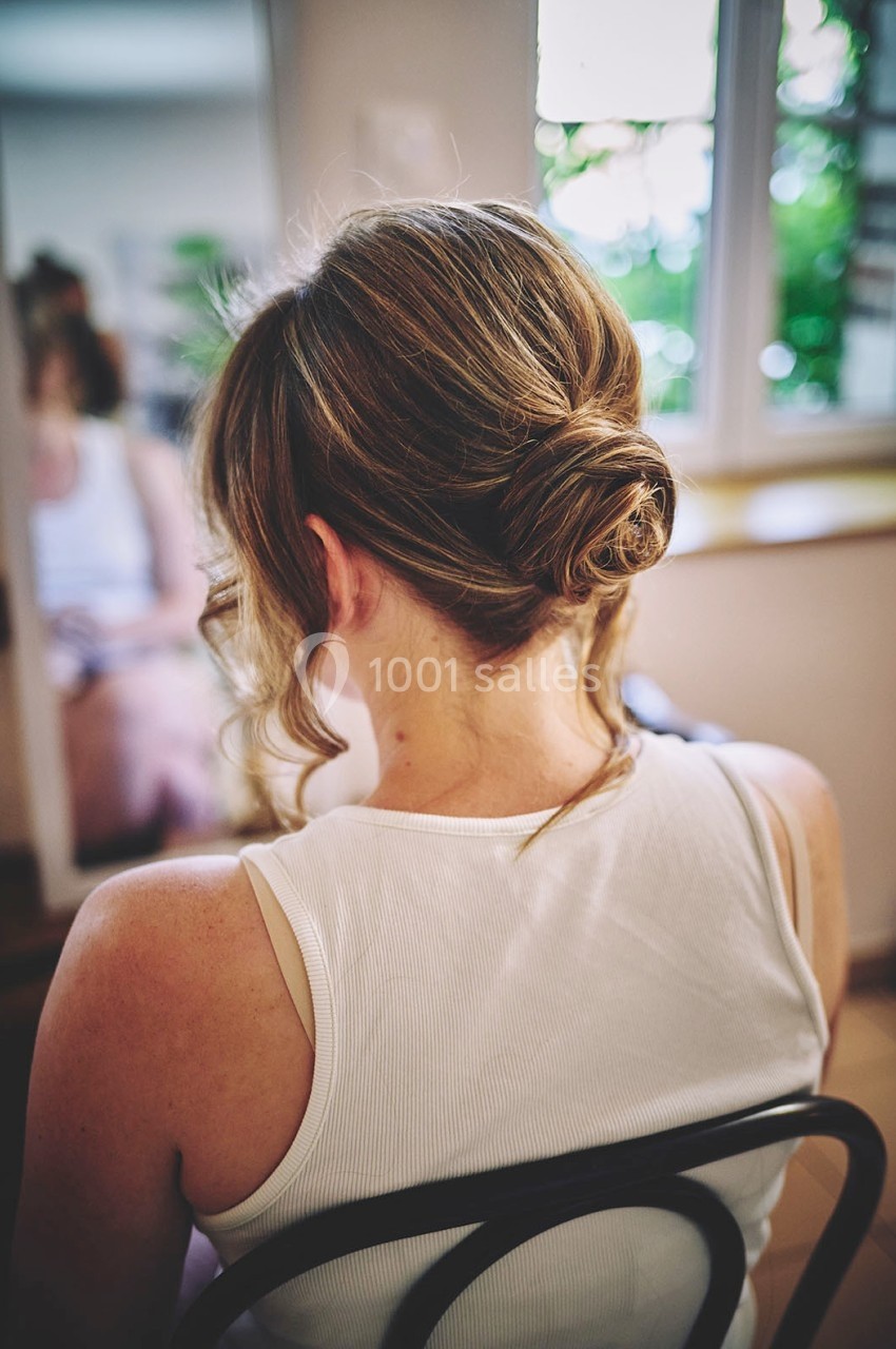Femme de dos avec un chignon élégant, assise sur une chaise devant un miroir dans une pièce lumineuse.