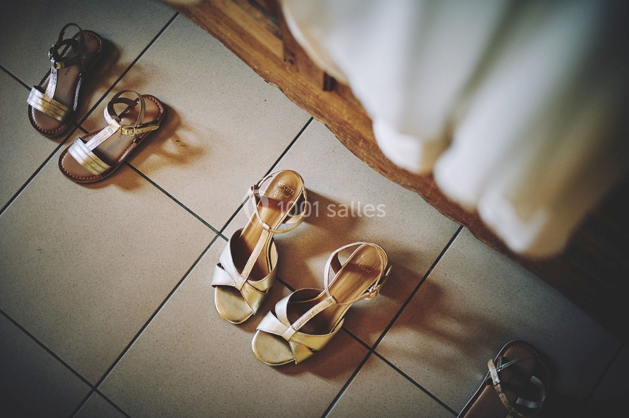 Sandales dorées et marron posées sur un sol carrelé beige, près d'une robe blanche suspendue.