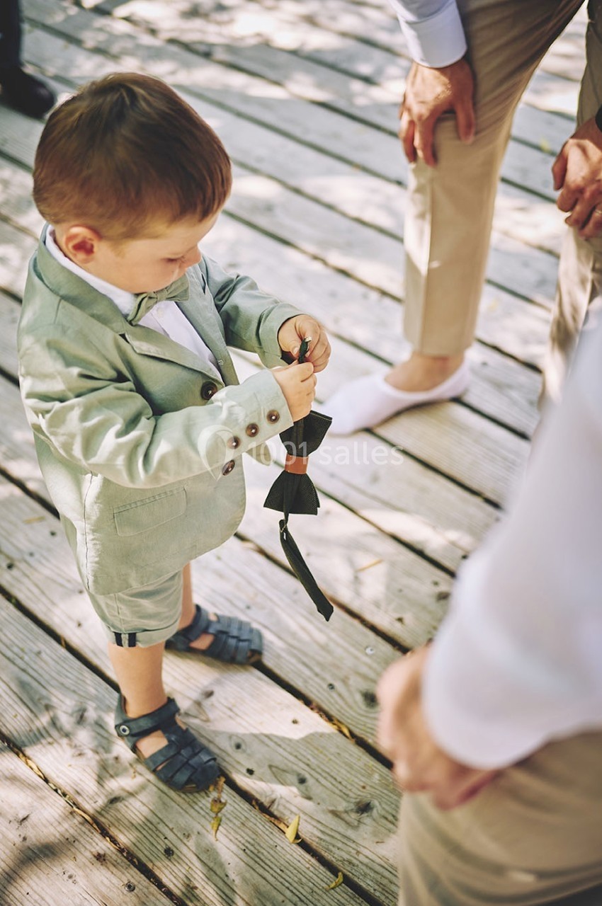 Un enfant en costume examine un nœud papillon noir sur une terrasse en bois, entouré d'adultes.