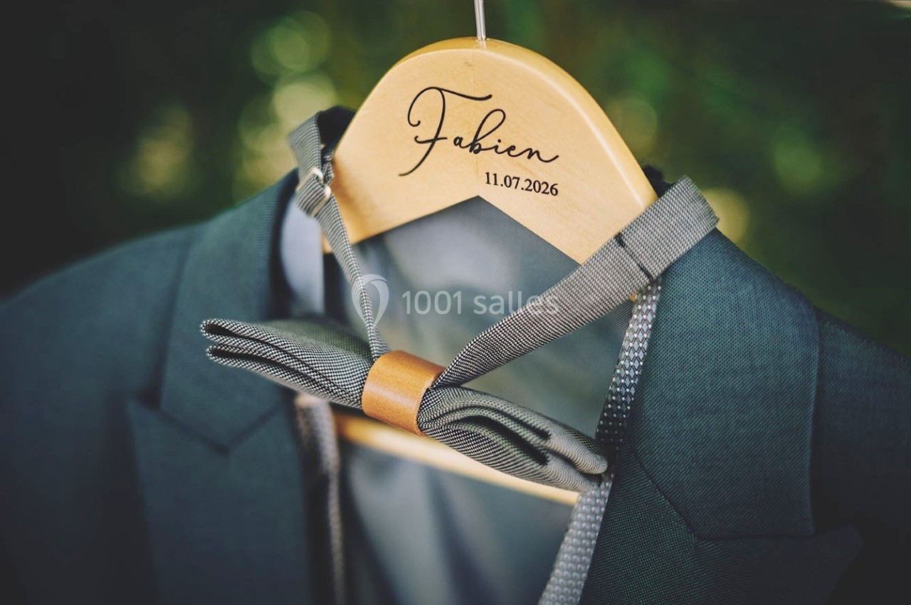 Un costume vert avec un nœud papillon gris suspendu à un cintre en bois gravé du prénom ’Fabien’ et d'une date.