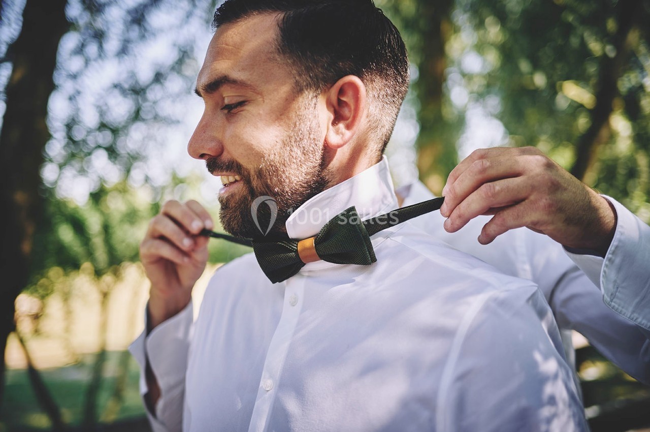 Un homme en chemise blanche ajuste un nœud papillon vert avec l'aide d'une autre personne dans un cadre extérieur verdoyant.