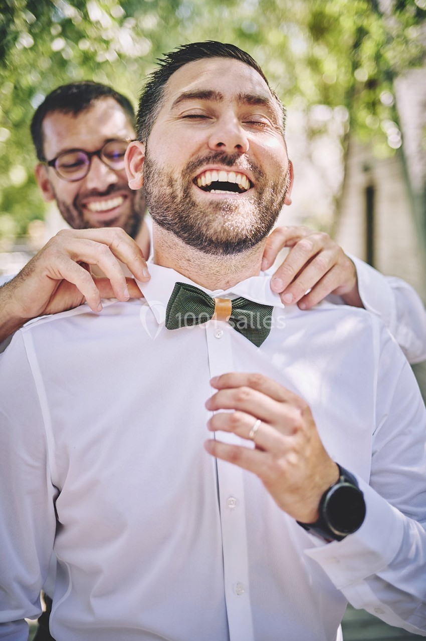 Deux hommes souriants, l'un ajustant le nœud papillon vert de l'autre, en extérieur sous un arbre.
