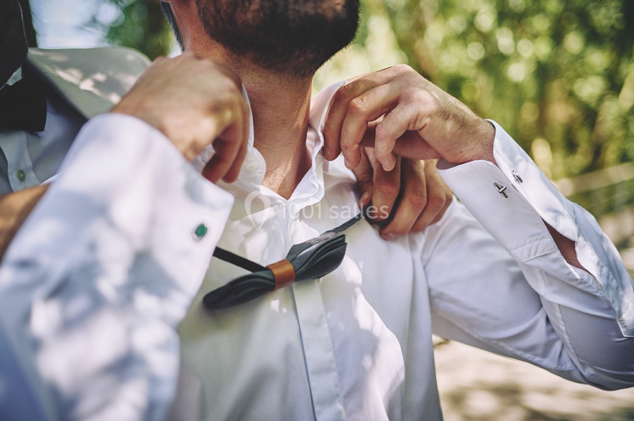 Un homme ajuste son nœud papillon noir sur une chemise blanche, aidé par une autre personne en arrière-plan flou.