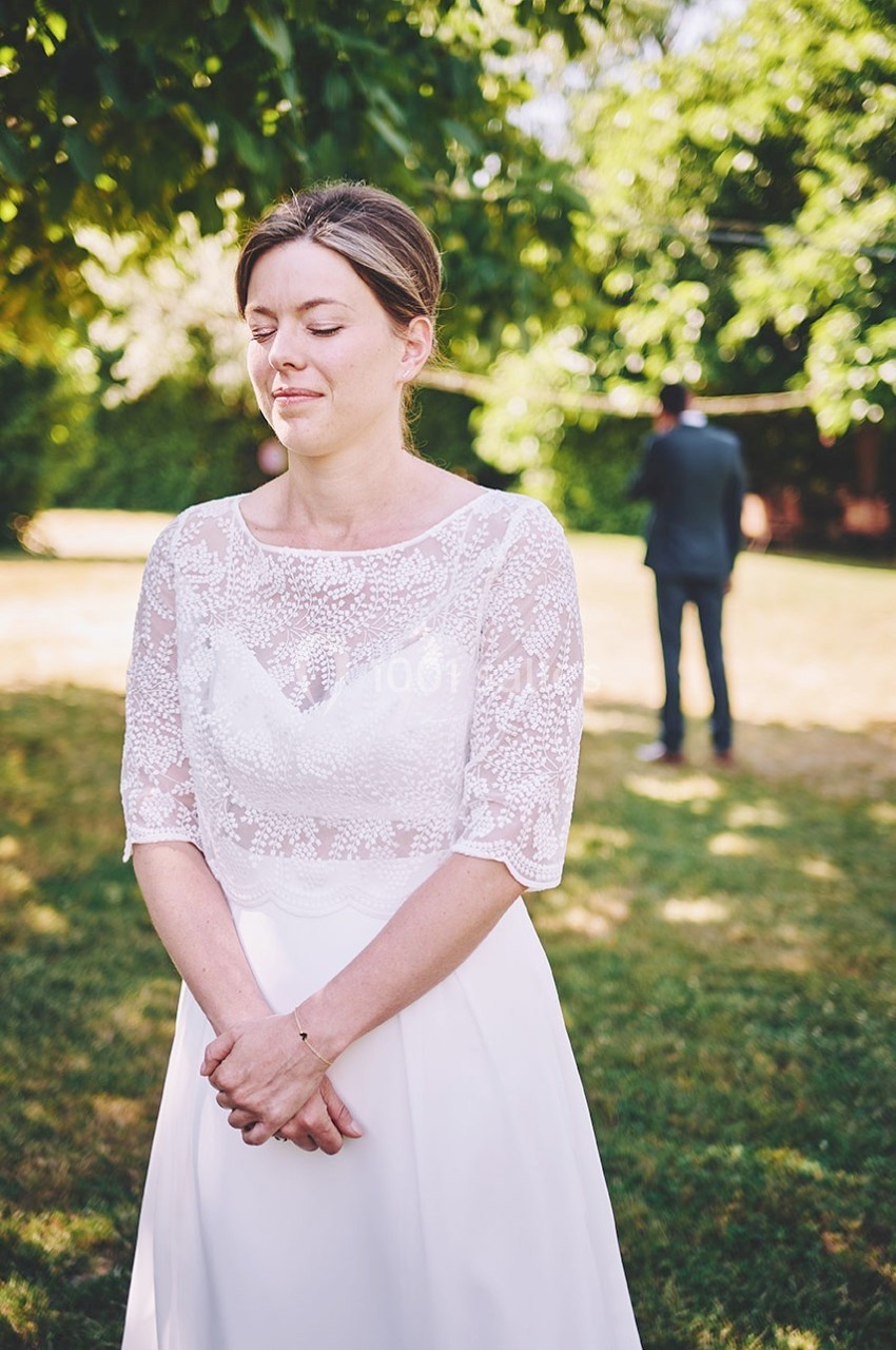 Une femme en robe blanche, les yeux fermés, debout dans un jardin ensoleillé, un homme flou en arrière-plan.
