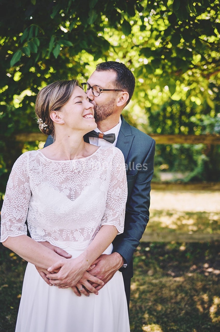 Un couple en tenue de mariage pose dans un jardin ensoleillé, entouré de verdure.