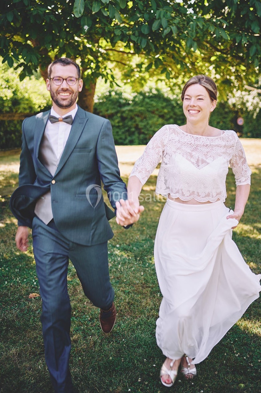 Un couple souriant en tenue de mariage marche main dans la main dans un jardin verdoyant.