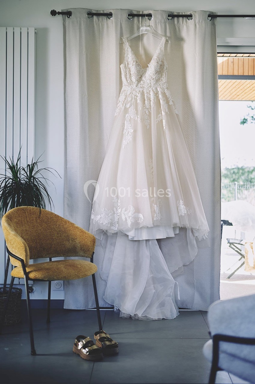 Robe de mariée blanche en dentelle suspendue devant un rideau beige, avec une chaise et des chaussures dorées au sol.