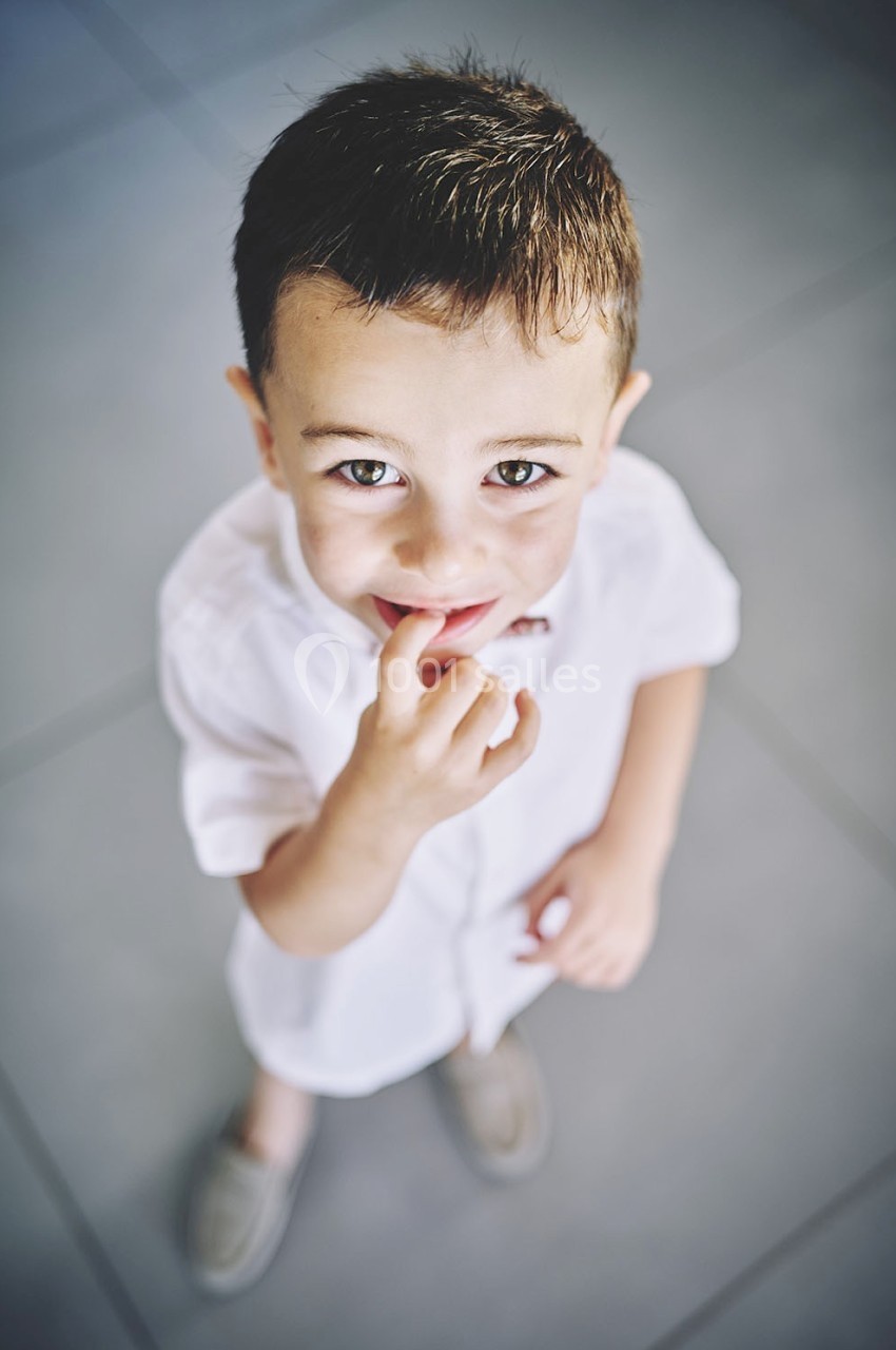 Un jeune garçon en chemise blanche regarde vers le haut en souriant, debout sur un sol carrelé gris.