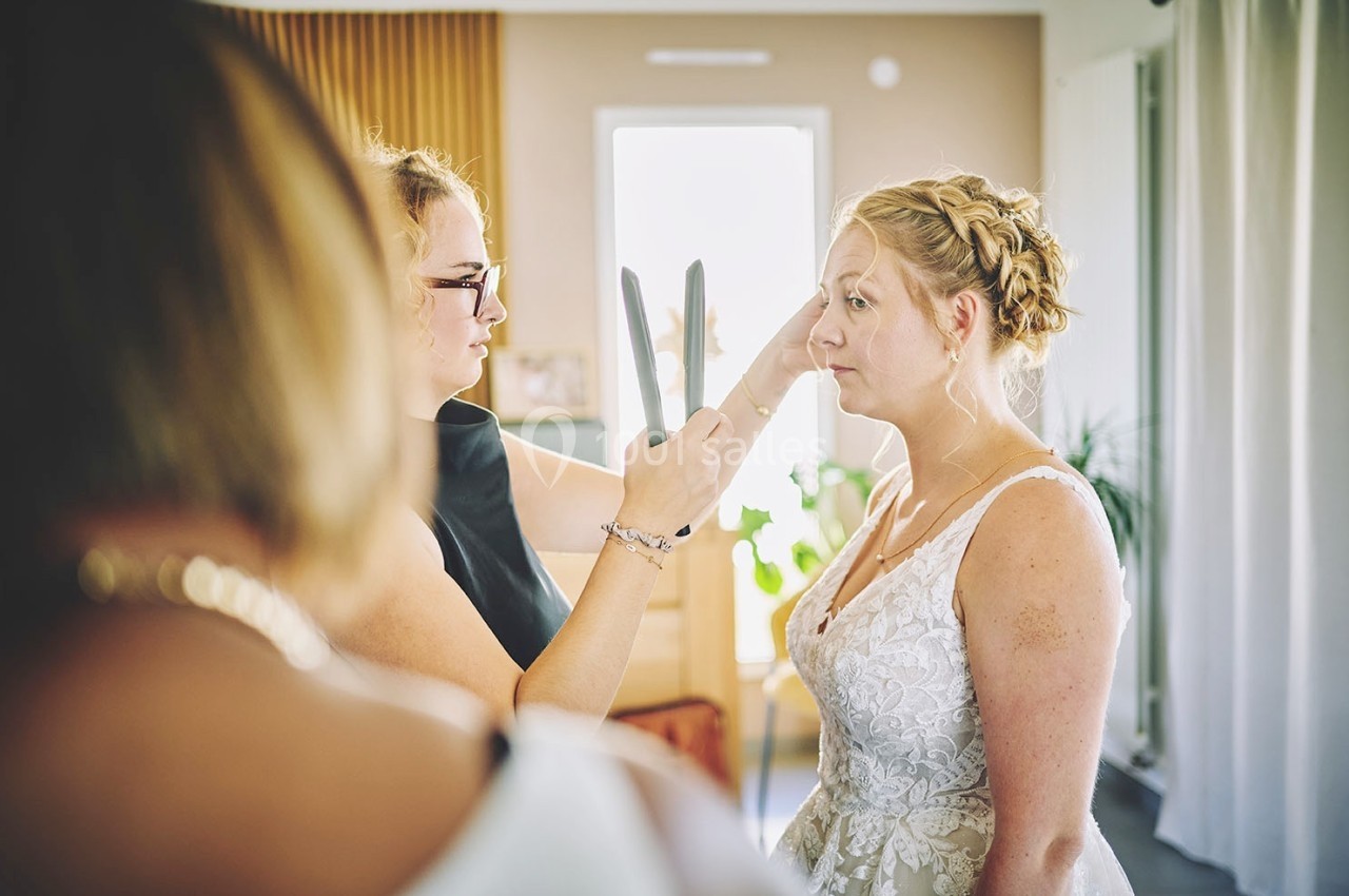 Une maquilleuse ajuste le maquillage d'une mariée dans une pièce lumineuse avant une cérémonie.