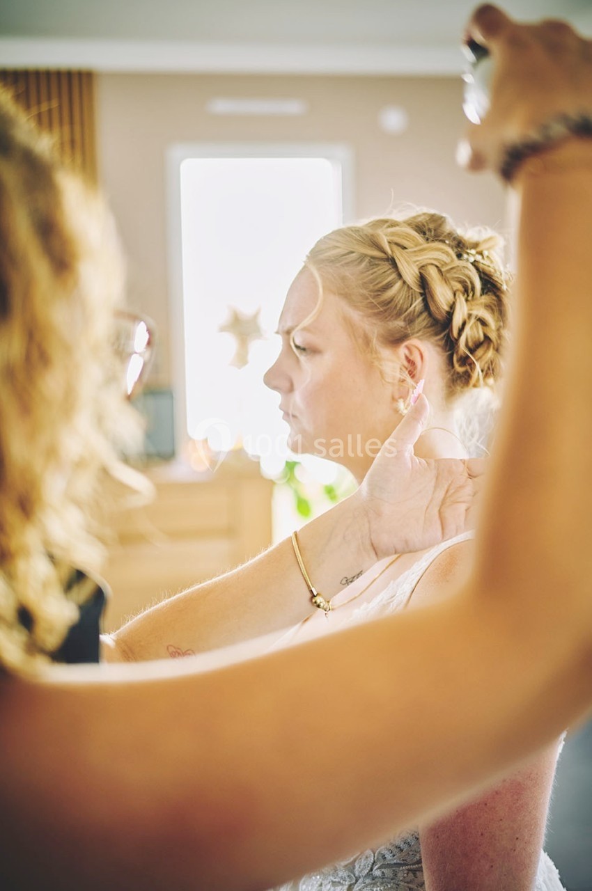 Une coiffeuse applique un spray sur les cheveux d'une femme coiffée d'une tresse, dans un intérieur lumineux.