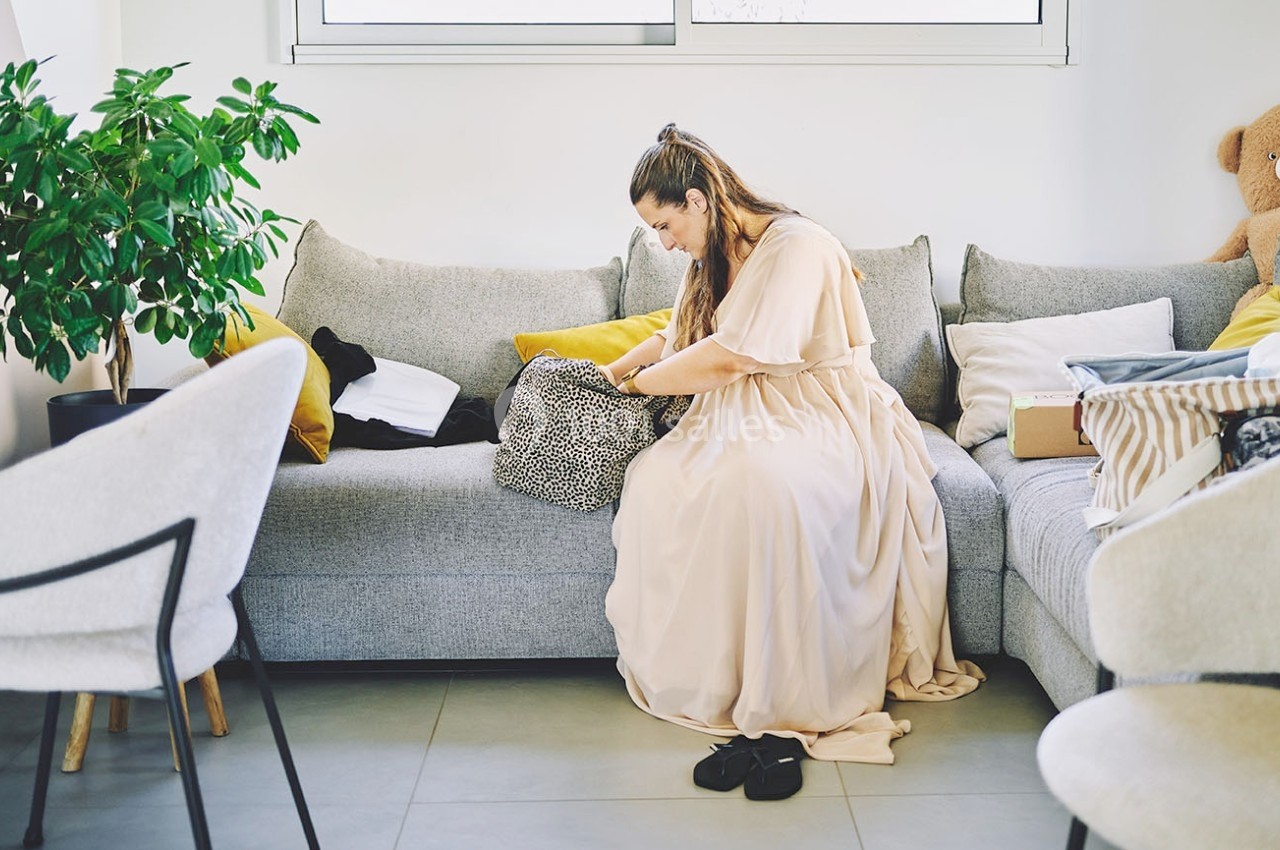 Une femme assise sur un canapé beige fouille dans un sac à motifs, entourée de coussins et d'objets divers.