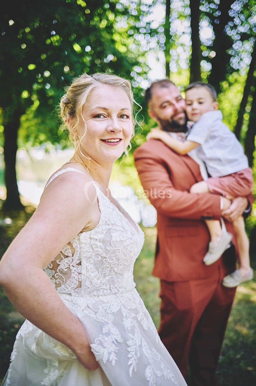 Une femme en robe de mariée sourit, avec un homme tenant un enfant en arrière-plan dans un cadre boisé.