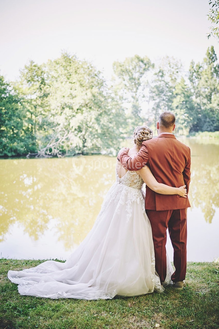 Un couple en tenue de mariage regarde un étang entouré d'arbres par une journée ensoleillée.