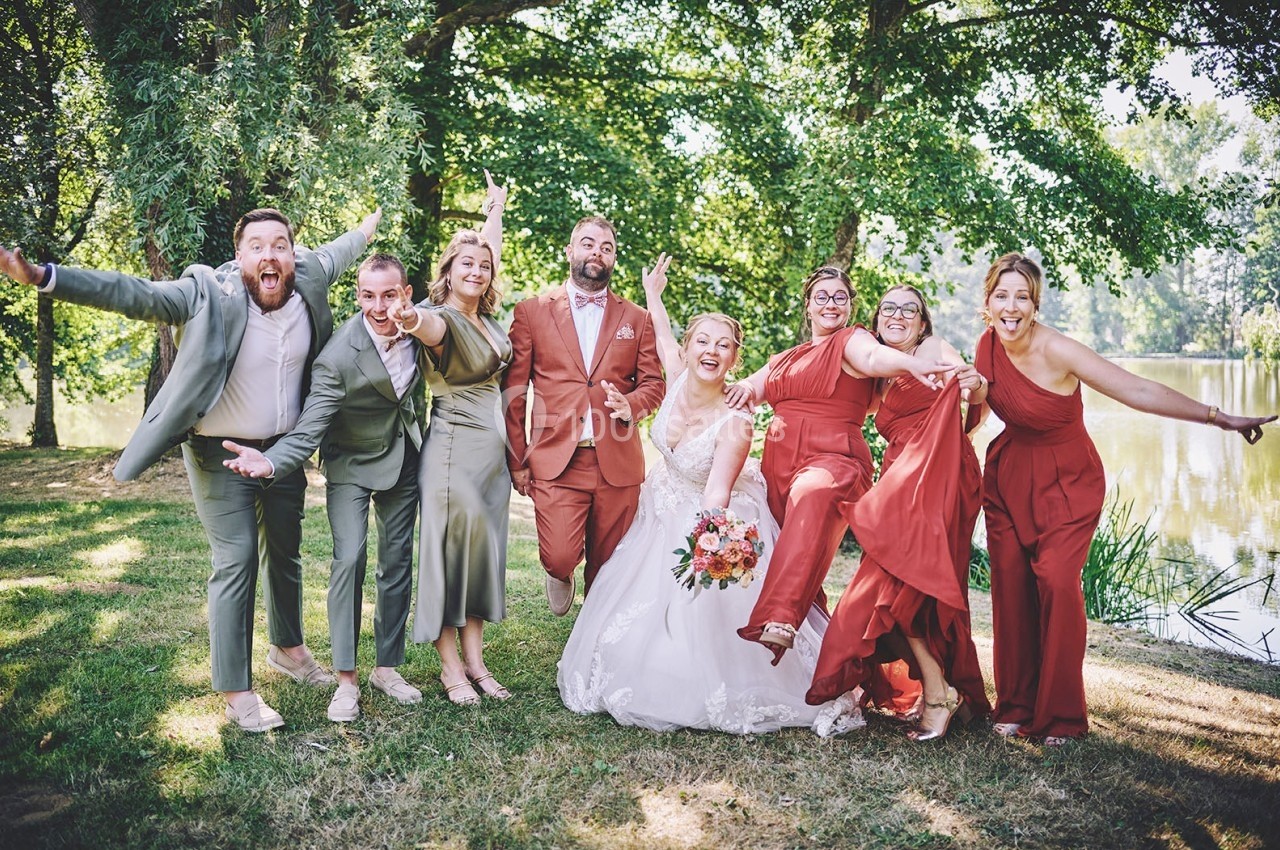 Un groupe joyeux pose en extérieur près d'un étang, avec une mariée au centre en robe blanche.