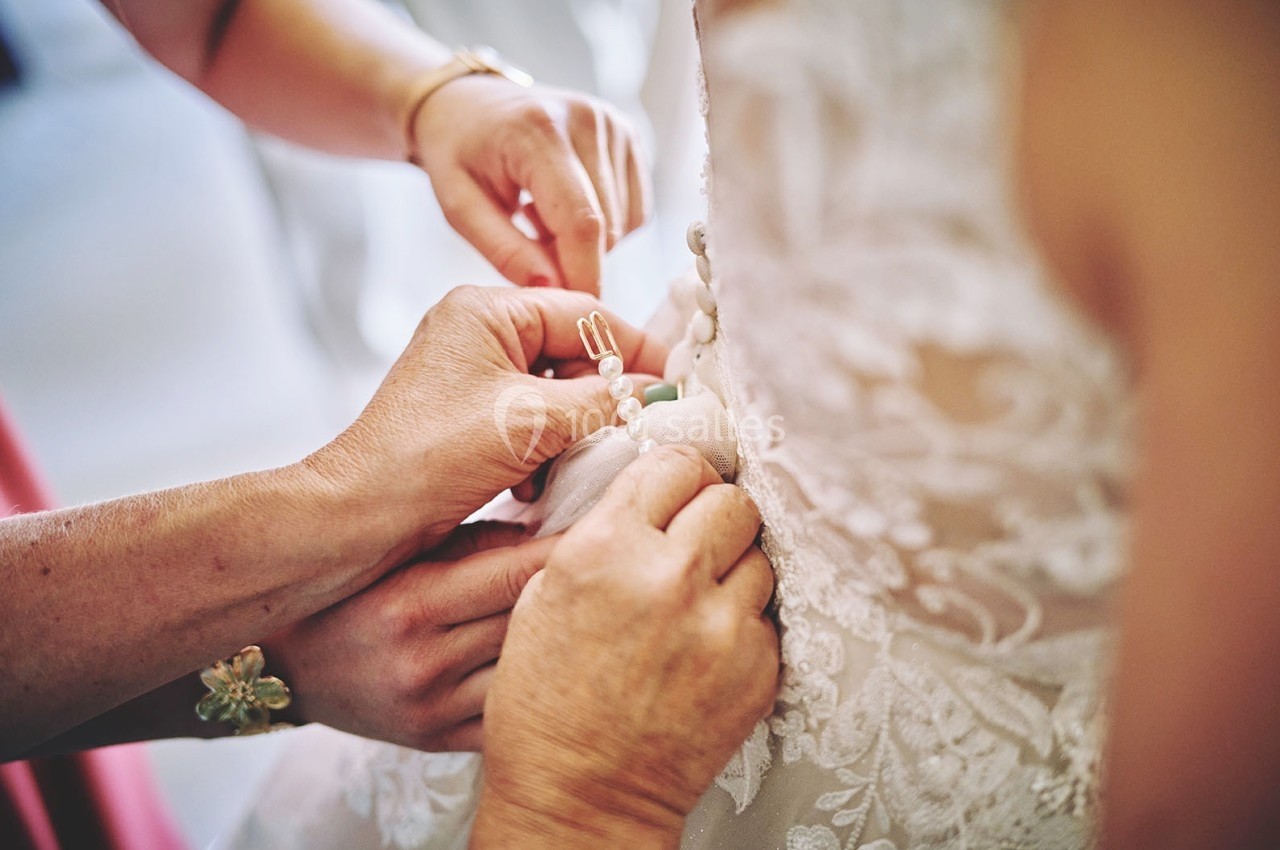 Des mains ajustent les boutons d'une robe de mariée en dentelle, mettant en valeur des détails délicats.