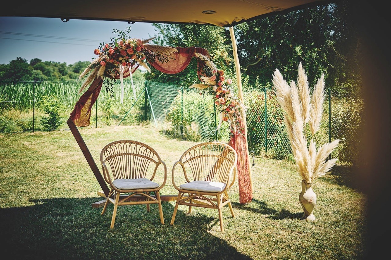 Arche de mariage décorée de fleurs et de tissu rose, avec deux chaises en rotin sur une pelouse en extérieur.