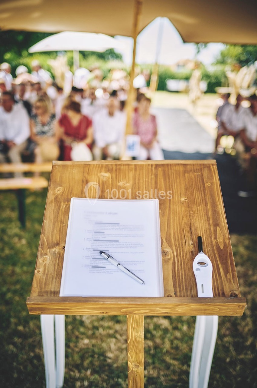 Pupitre en bois avec documents, stylo et micro, devant un public assis en extérieur sous des parasols.
