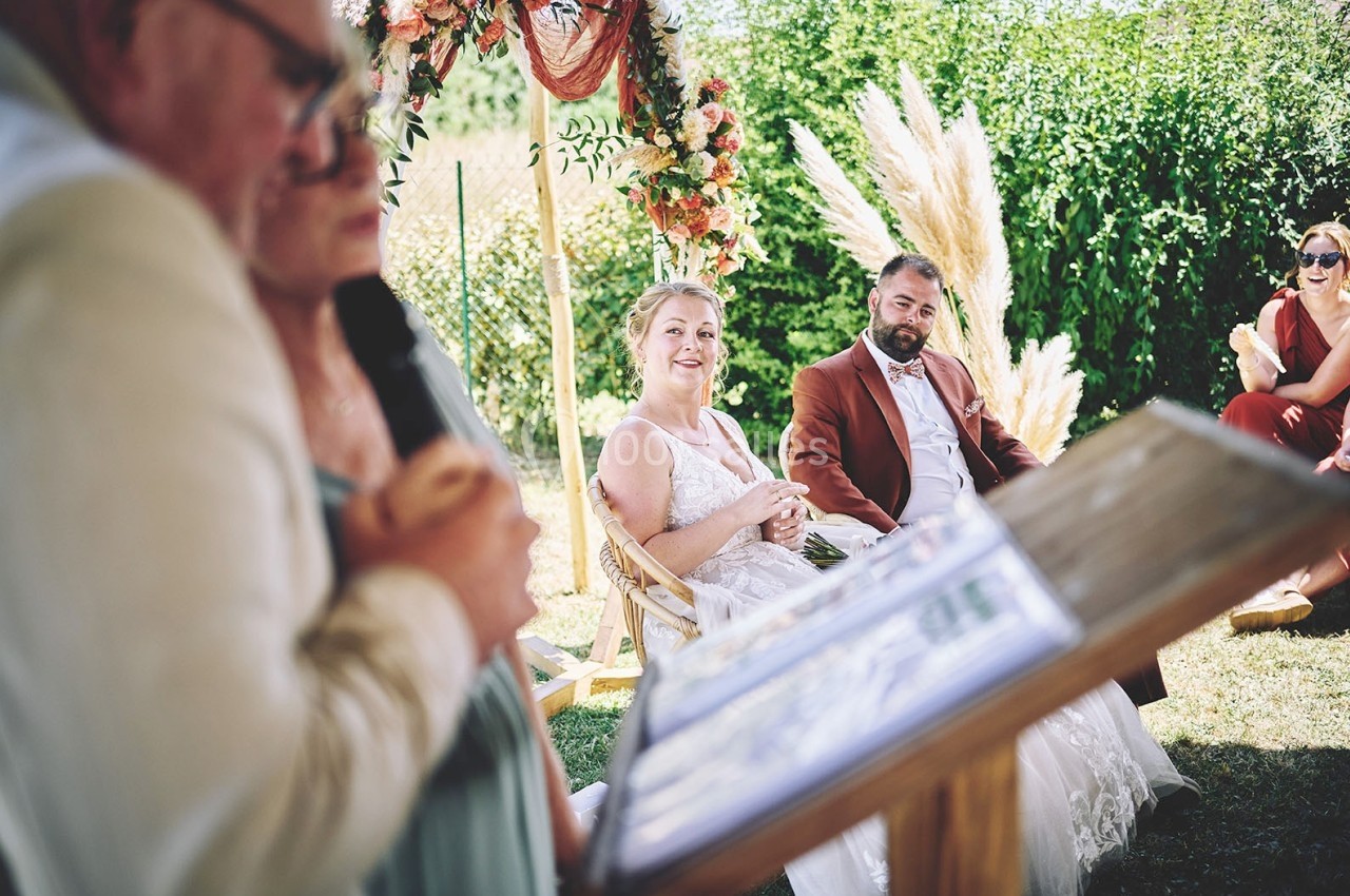 Un couple assis en extérieur lors d'une cérémonie, entouré de verdure et d'une arche florale.