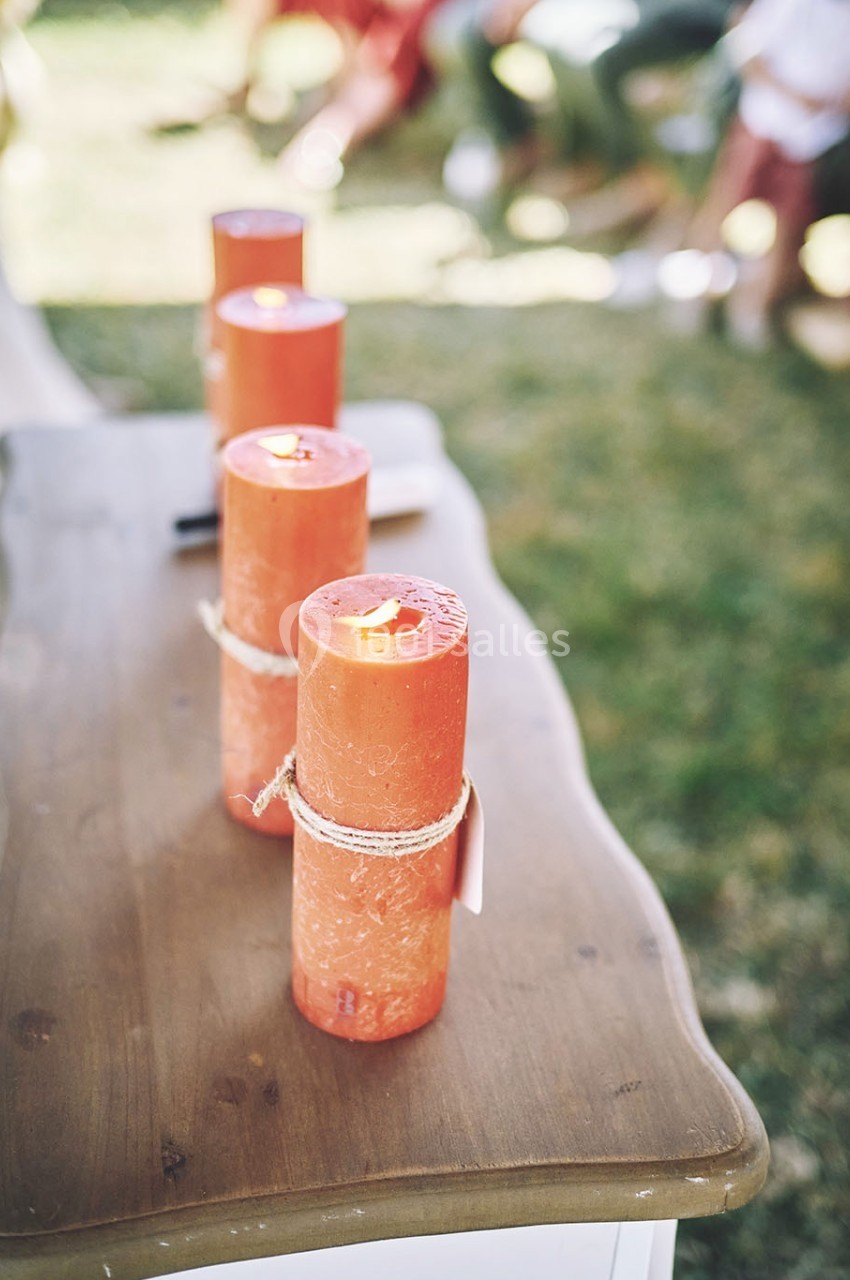 Quatre bougies orange allumées, attachées avec de la ficelle, posées sur une table en bois dans un environnement extérieur.