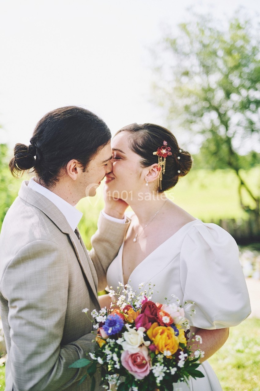 Un couple s'embrasse tendrement en extérieur, la mariée tenant un bouquet coloré, sous une lumière naturelle.