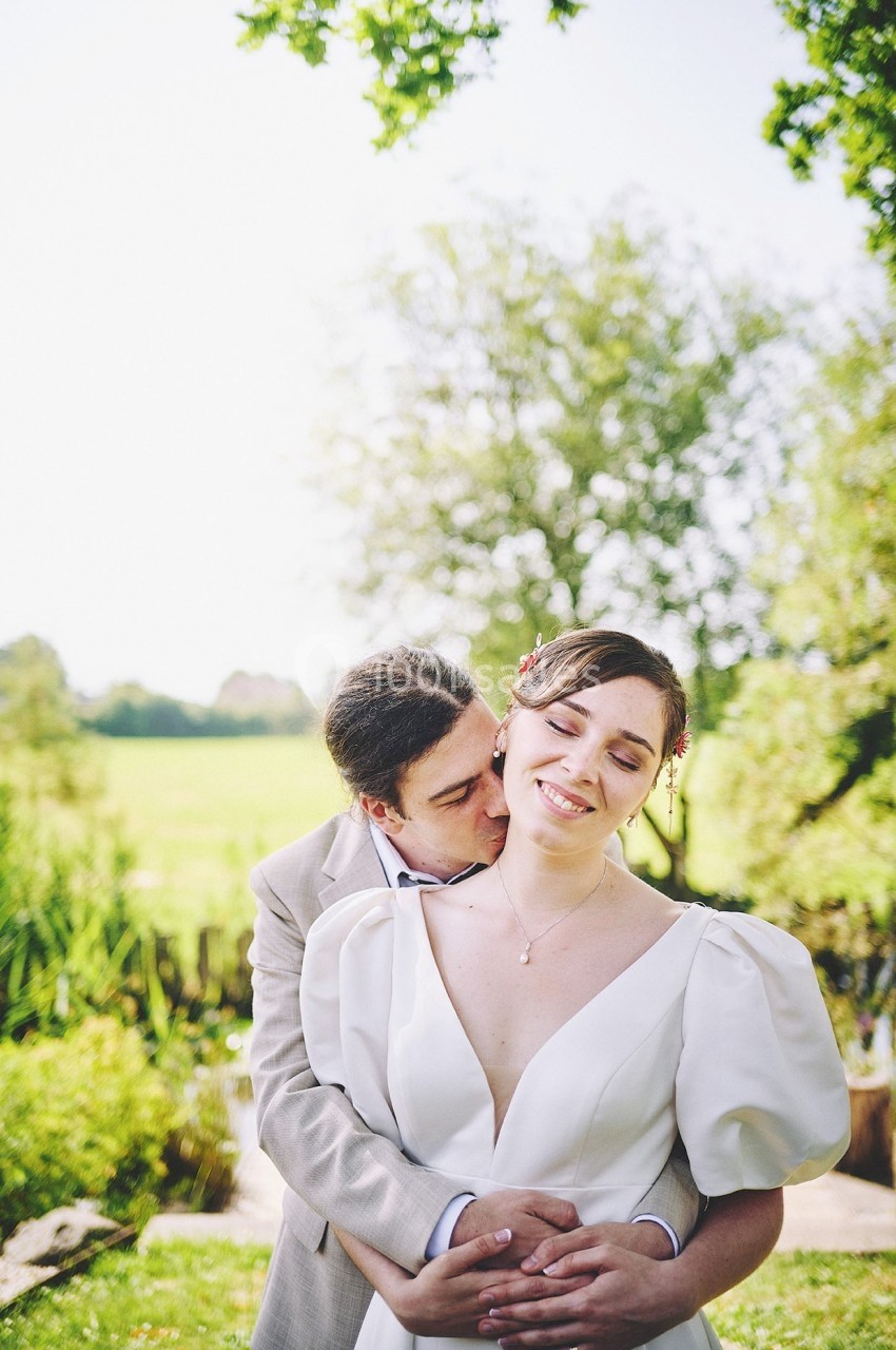 Un couple en tenue de mariage pose dans un jardin verdoyant sous un ciel clair.