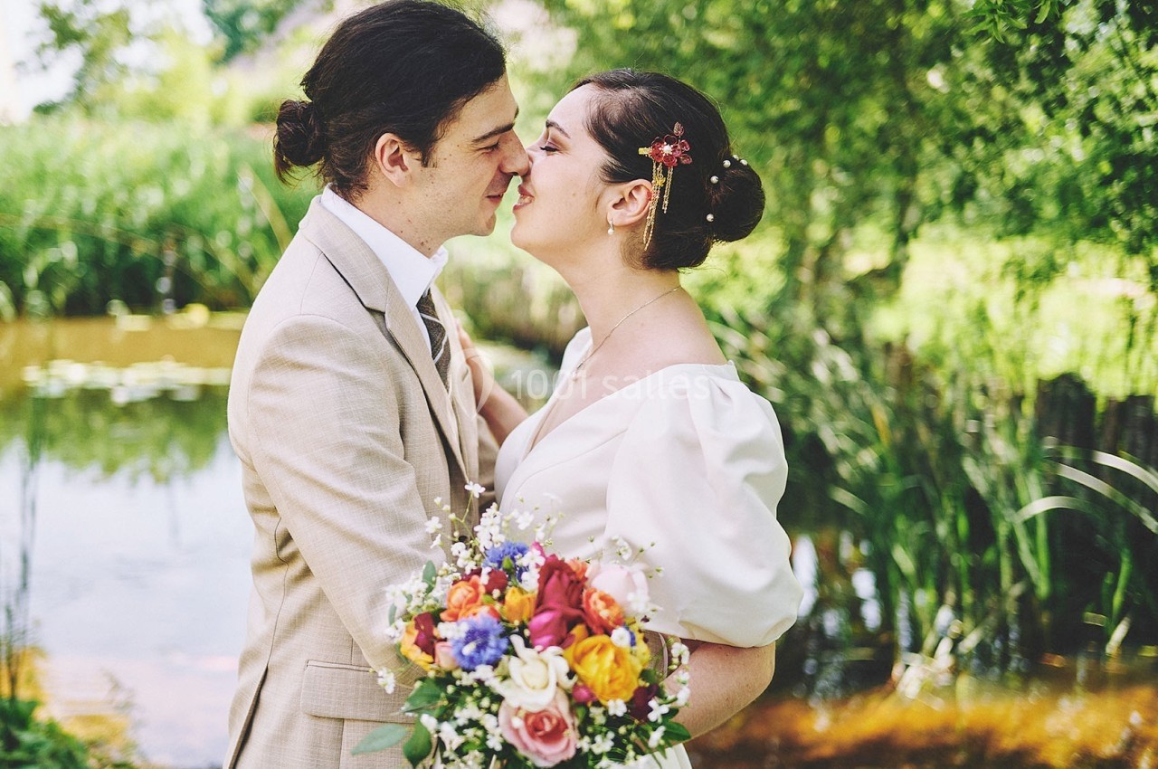 Un couple de mariés s'embrasse tendrement près d'un étang, entouré de verdure, la mariée tenant un bouquet coloré.