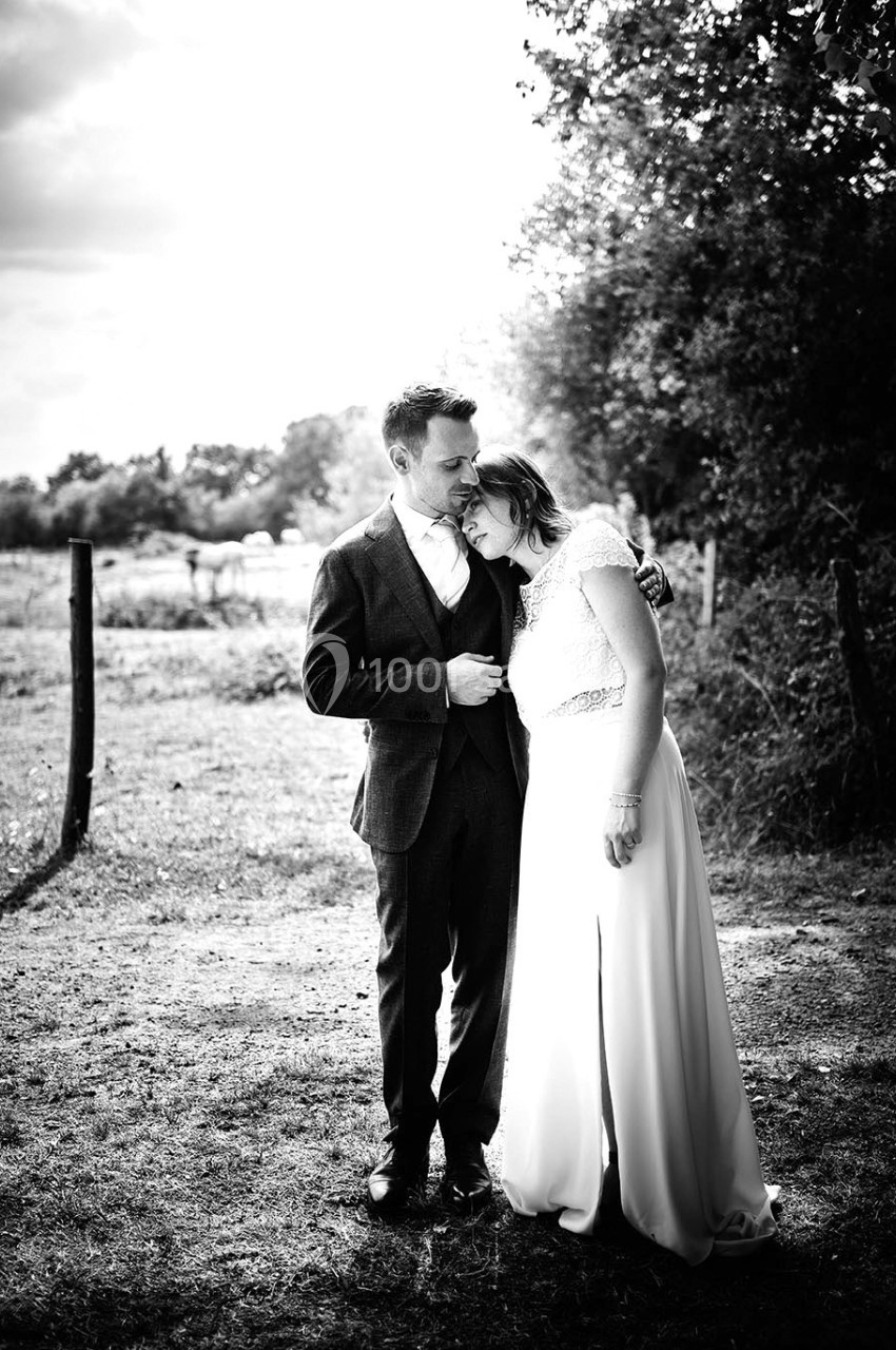 Un couple en tenue de mariage se tient tendrement dans un paysage champêtre en noir et blanc.