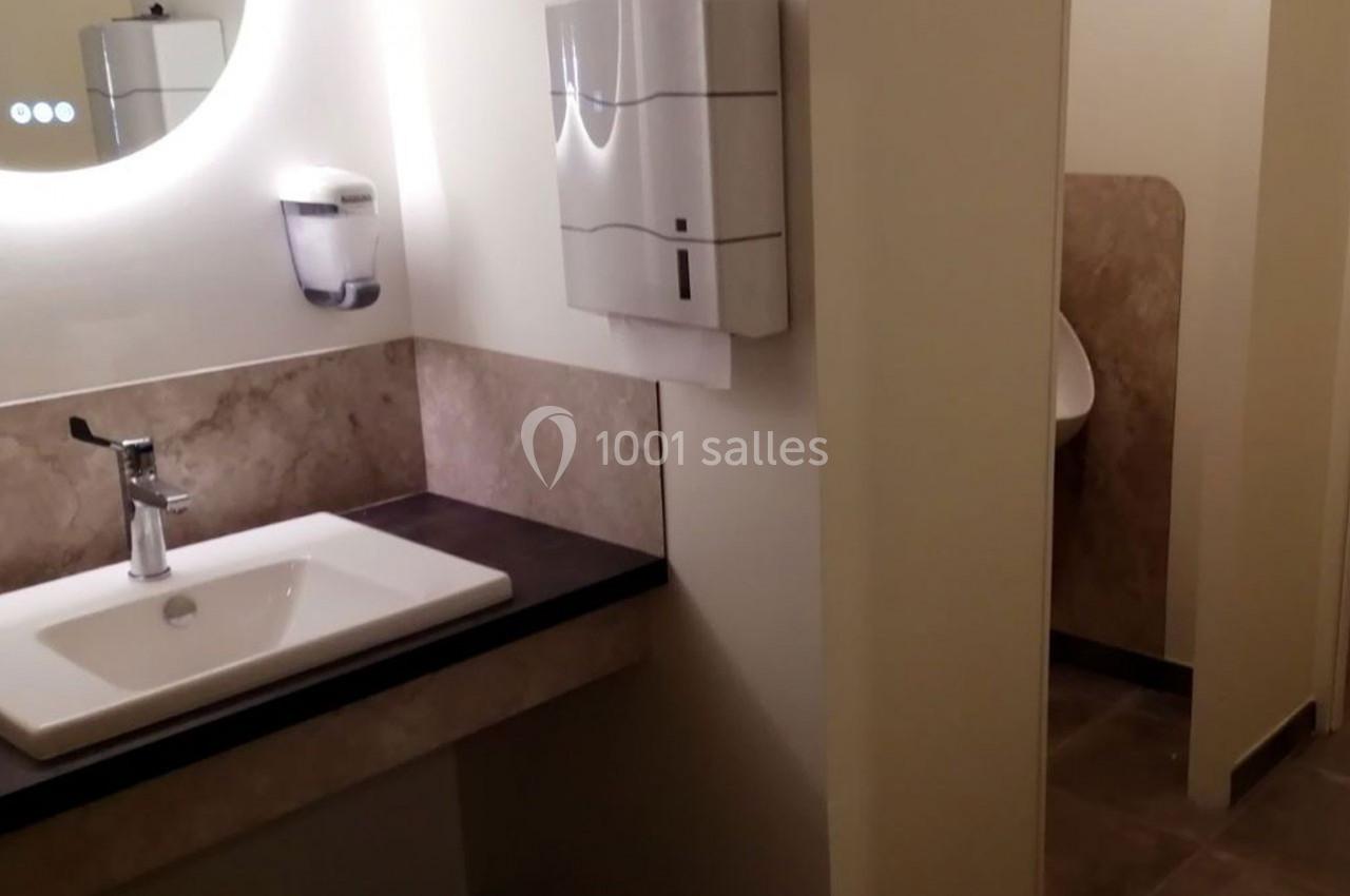 Lavabo avec miroir éclairé, distributeur de savon et essuie-mains, adjacent à une cloison séparant un urinoir.