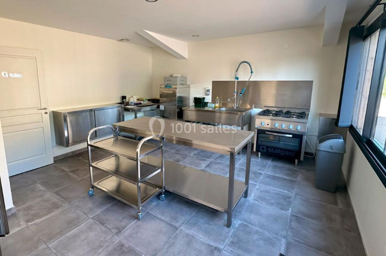 Cuisine professionnelle équipée avec plans de travail en inox, évier, cuisinière, étagères et éclairage naturel.