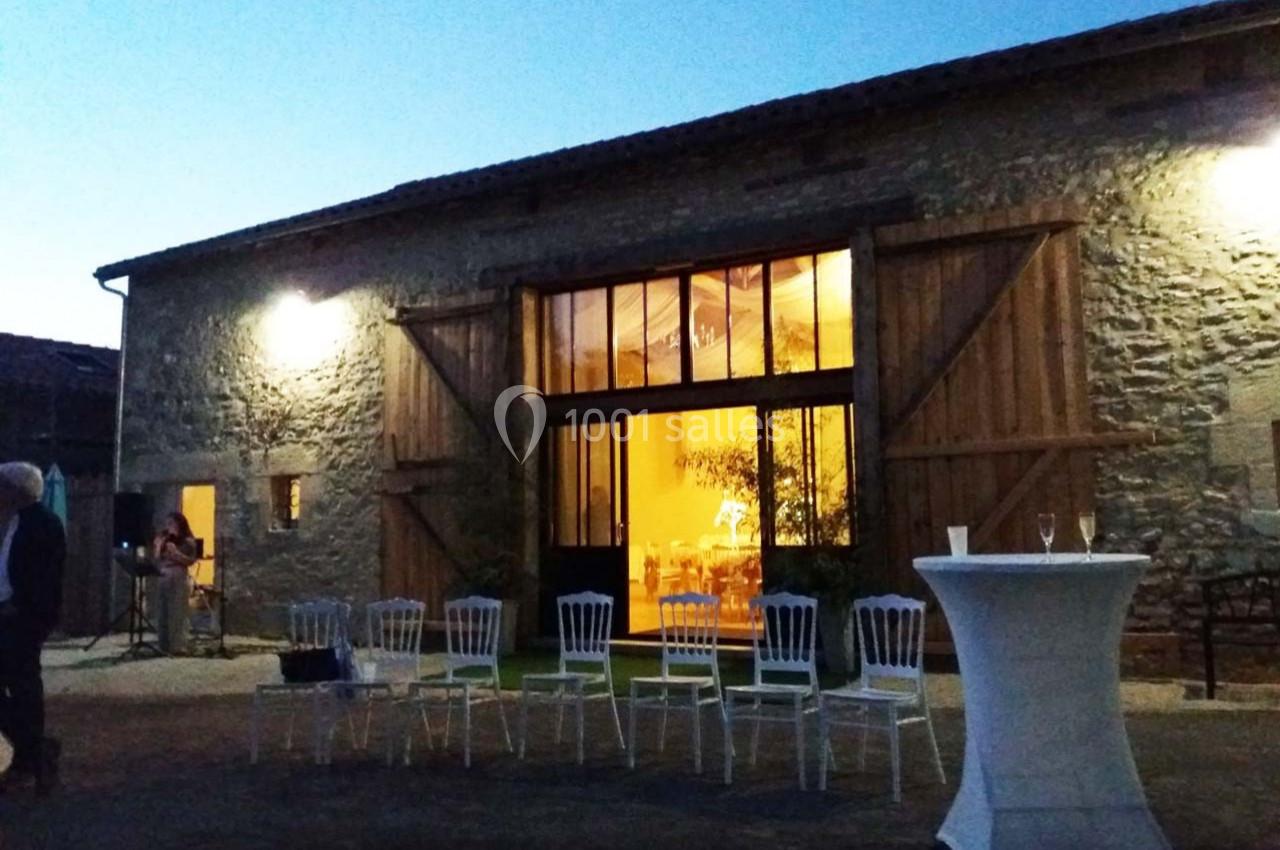 Façade d'une grange rénovée avec portes en bois ouvertes, éclairée de l'intérieur, chaises et table haute à l'extérieur.