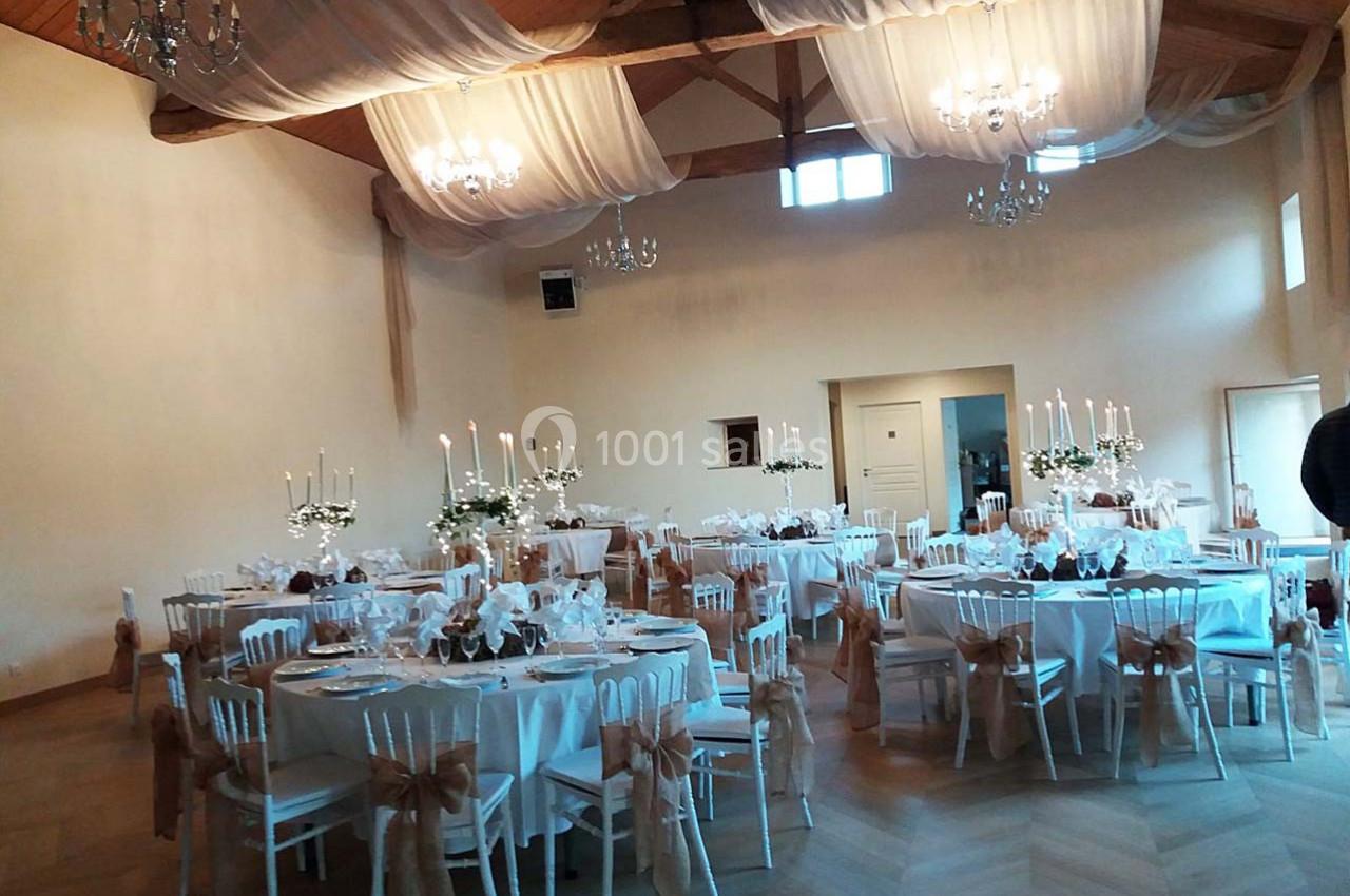 Salle de réception décorée avec des tables rondes, nappes blanches, chaises ornées de rubans et chandeliers.