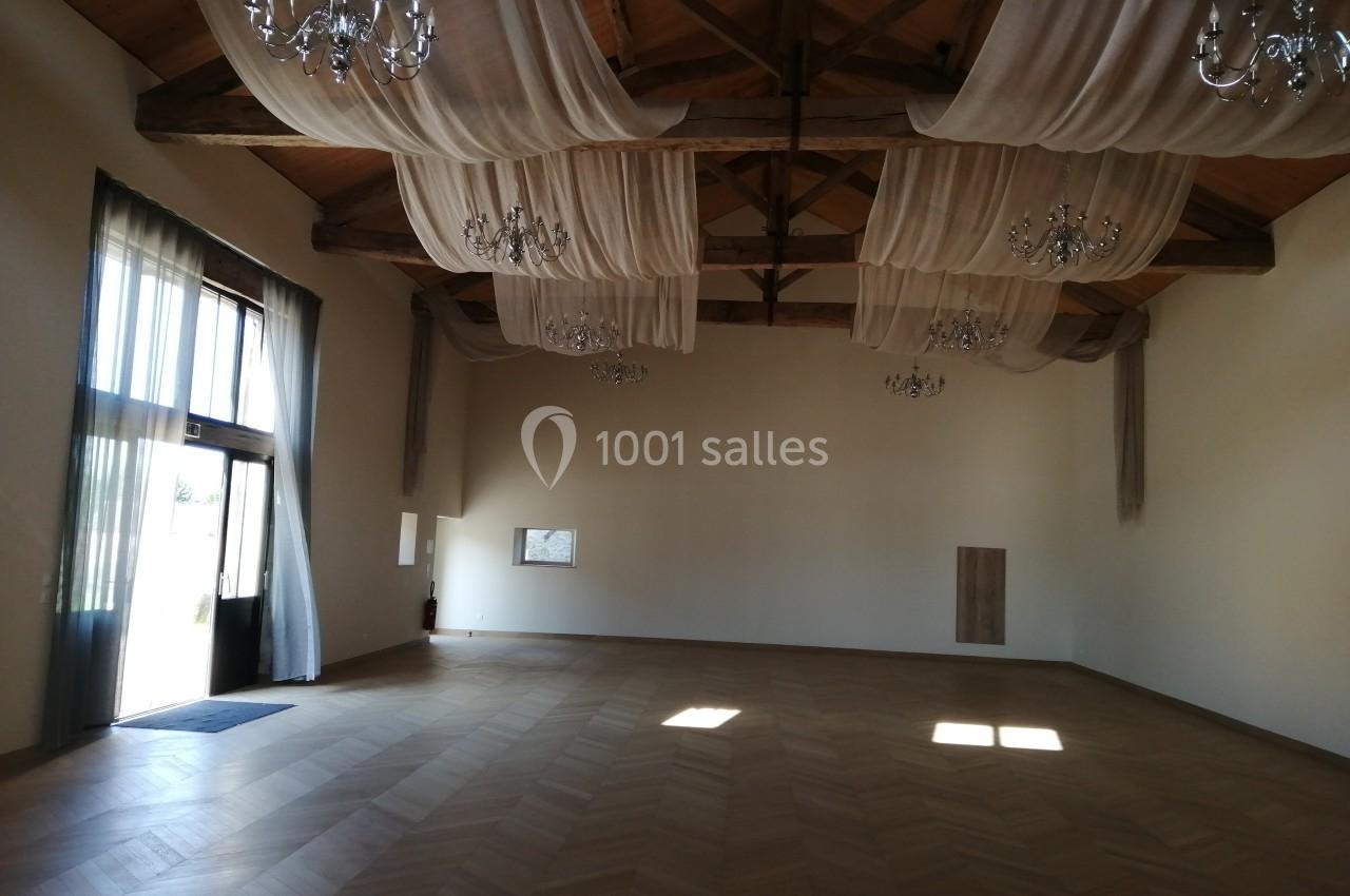 Salle spacieuse avec parquet en bois, poutres apparentes, lustres et rideaux suspendus, éclairée par des fenêtres.