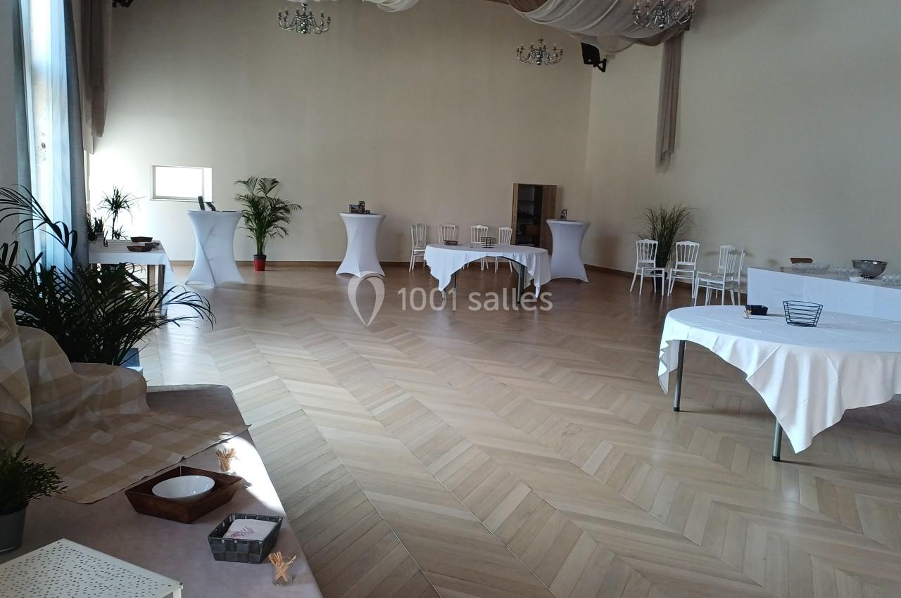 Salle lumineuse avec parquet, tables rondes et hautes décorées, plantes et nappes blanches, prête pour un événement.