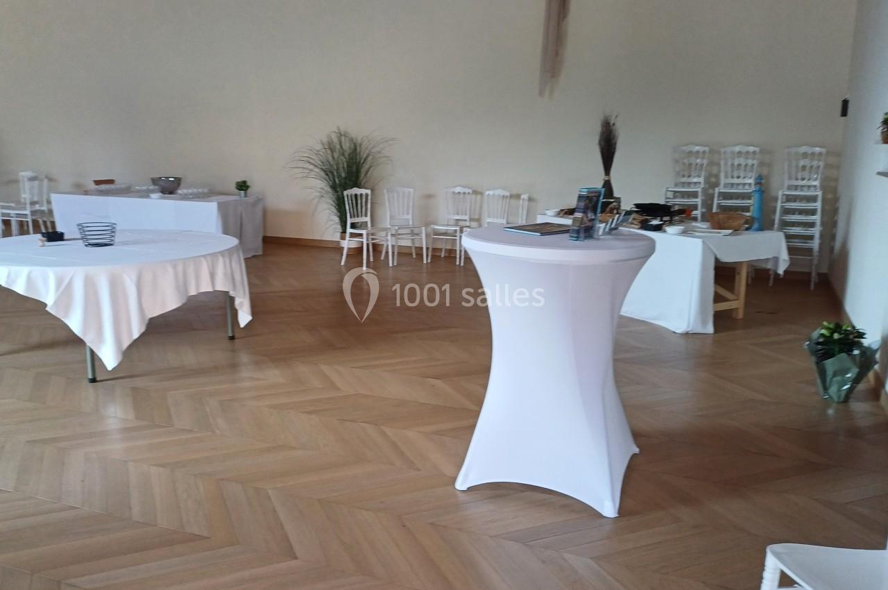 Salle lumineuse avec parquet, tables dressées avec nappes blanches, chaises empilées et décorations végétales.