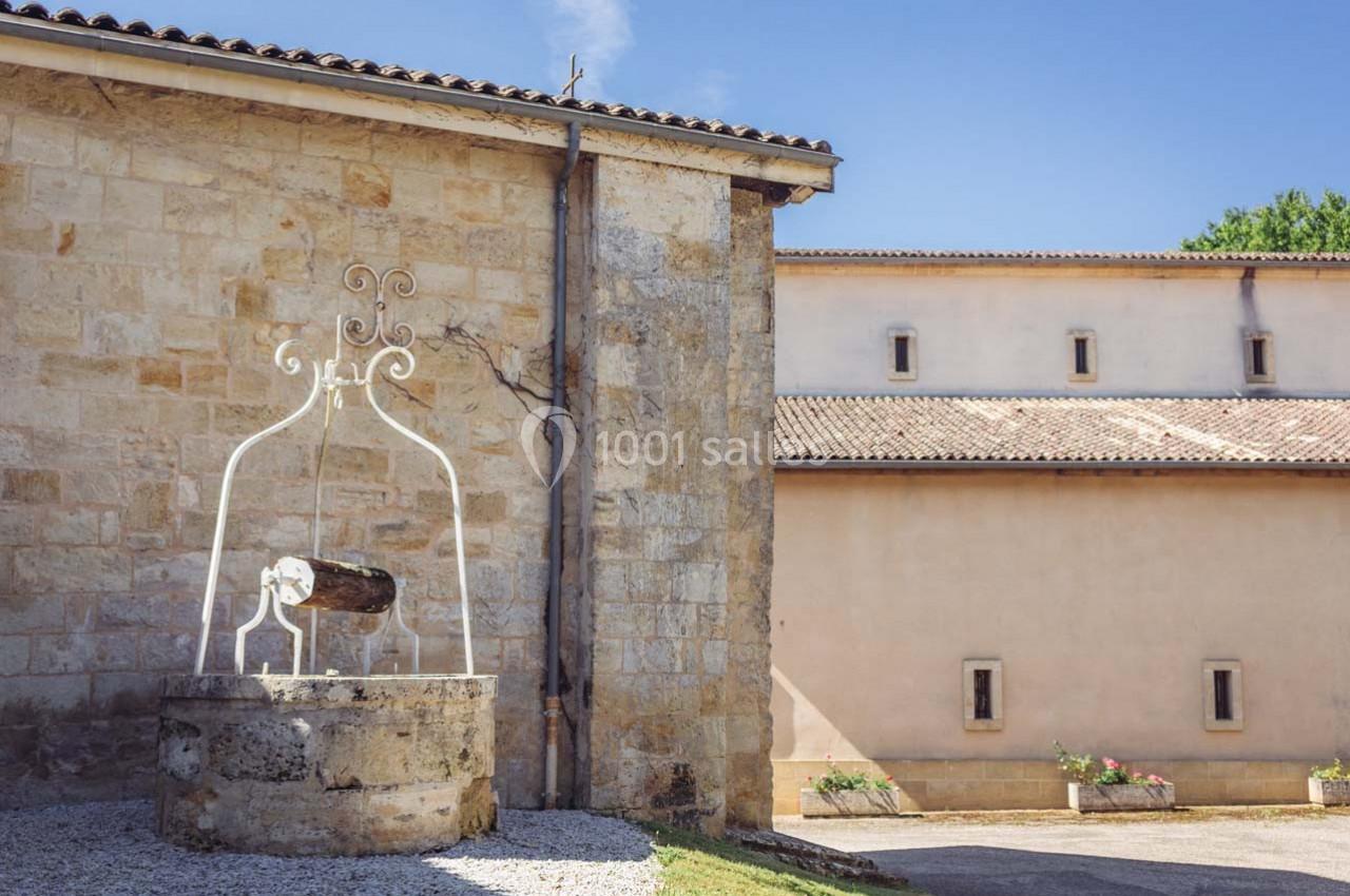 Un puits en pierre avec une structure métallique décorative, situé près d'un bâtiment en pierre sous un ciel dégagé.