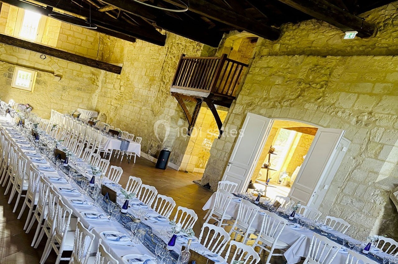 Grande salle en pierre avec de longues tables dressées pour un repas, chaises blanches et mezzanine en bois visible.