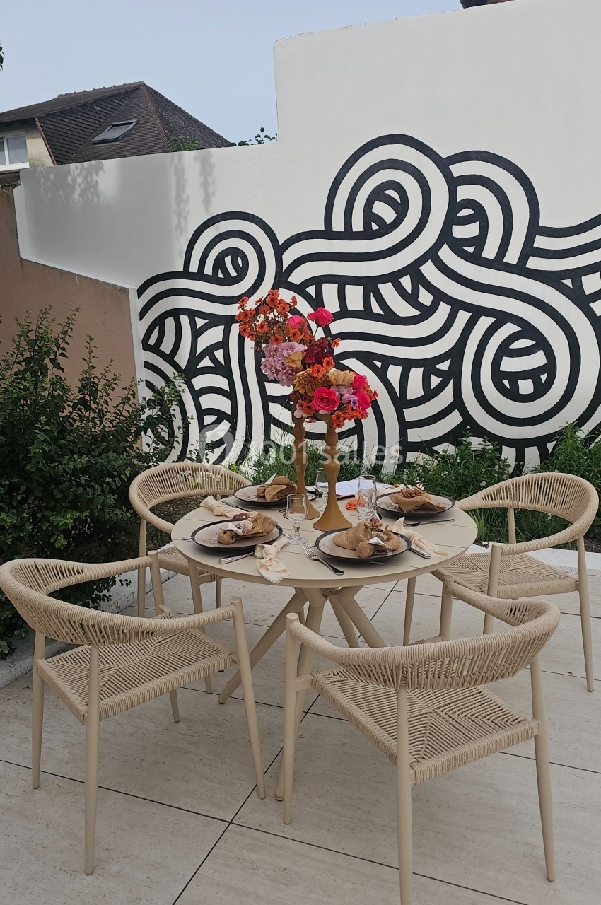 Table de jardin beige dressée pour un repas, entourée de chaises en corde, avec un bouquet coloré et une fresque murale en…