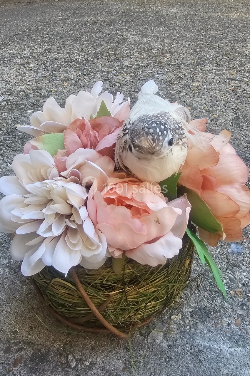 Un petit oiseau perché sur un nid décoré de fleurs roses et blanches, posé sur un sol en béton.